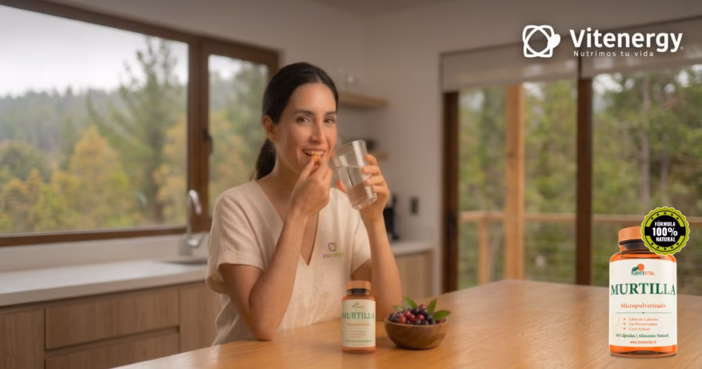 Una mujer sonríe mientras consume una cápsula de murtilla FuenteVital con un vaso de agua, sentada en una cocina iluminada. Un frasco de suplemento FuenteVital Murtilla Micropulverizado y un tazón de murtillas frescas están en la mesa. A la izquierda, logo de Vitenergy; a la derecha, título: 'Beneficios de la Murtilla'.