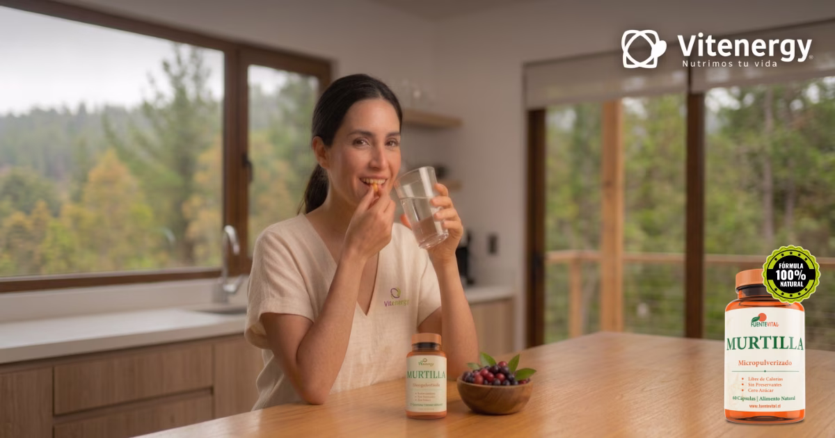 Una mujer sonríe mientras consume una cápsula de murtilla FuenteVital con un vaso de agua, sentada en una cocina iluminada. Un frasco de suplemento FuenteVital Murtilla Micropulverizado y un tazón de murtillas frescas están en la mesa. A la izquierda, logo de Vitenergy; a la derecha, título: 'Beneficios de la Murtilla'.